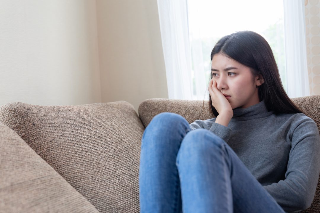 Unhappy Asian young woman siting alone on couch with feeling sadness.